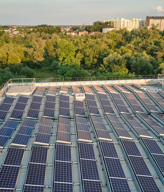 Rooftop solar panels installed on an industrial building, surrounded by trees and suburban landscape in the background.